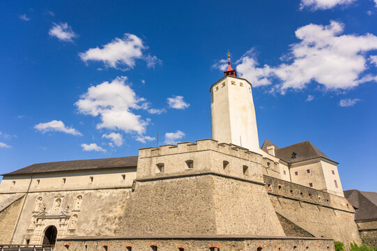 Esterhazy Castle located in Forchtenstein Austria showcases a massive stone fortress under a clear blue sky with scattered fluffy clouds illuminating the structure. The castle features tall towers