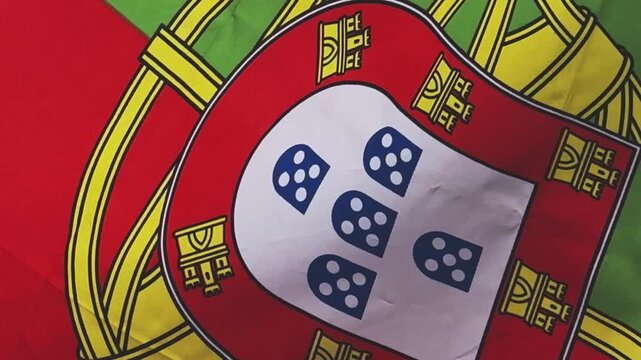 A textured close up of the Portuguese flag showing the central coat of arms and the vibrant green and red fabric rippling softly. The woven material is illuminated by natural light.