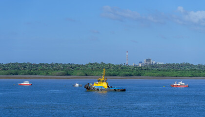 Fototapeta premium Vibrant yellow and blue tugboat navigates calm blue waters alongside smaller vessels. The background reveals a lush green shoreline contrasting with an industrial plant and tall smokestack
