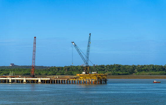 Industrial marine construction site featuring a heavy yellow pier structure with tall lattice boom cranes over blue water. A dense green coastal forest forms the background