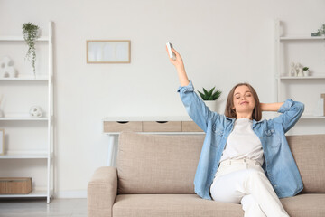Young woman relaxing on a comfortable sofa at home, adjusting the air conditioner with a remote control for comfort, She is enjoying a moment of peace and comfort at home