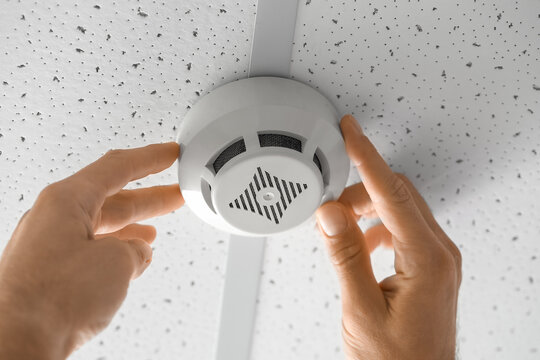 Man installing modern smoke detector on ceiling in room, closeup