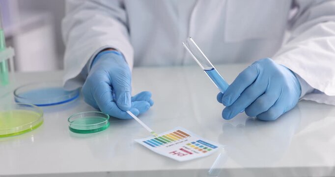 Scientist holding PH test strip and glass tube with liquid at white table in laboratory, closeup
