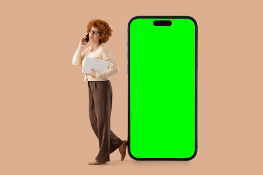 Young woman with laptop talking on phone next to large smartphone with green screen, digital communication and technology