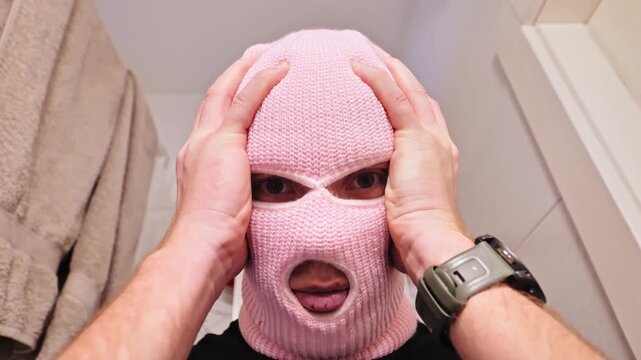 Person wearing pink balaclava pulling at fabric around face while looking into camera.