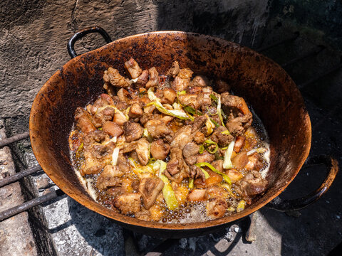 Hand stirs meat in a rusty wok over open flame. Fried chunks sizzle in oil, garlic, and greens. Dusty stone wall frames the rustic cooking scene. Hot air rises from the fire beneath the wok, Fritada