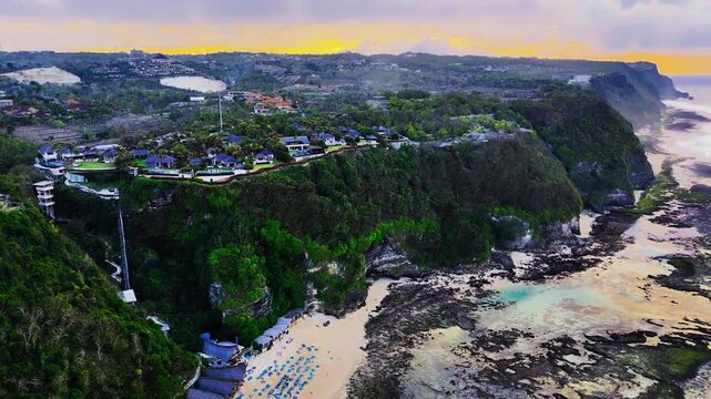 A smooth, high-altitude aerial pullback revealing the dramatic verticality of the Uluwatu cliffs and the rugged, rock-strewn coastal beach below during sunset.