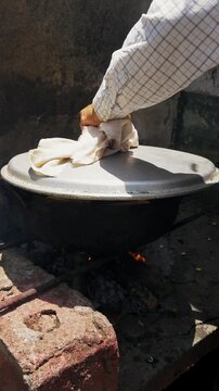 Hand stirs meat in a rusty wok over open flame. Fried chunks sizzle in oil, garlic, and greens. Dusty stone wall frames the rustic cooking scene. Hot air rises from the fire beneath the wok, Fritada