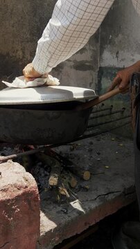 Hand stirs meat in a rusty wok over open flame. Fried chunks sizzle in oil, garlic, and greens. Dusty stone wall frames the rustic cooking scene. Hot air rises from the fire beneath the wok, Fritada