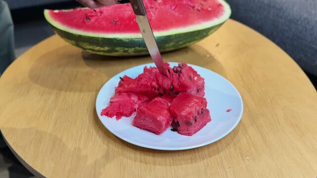 hot of fresh, juicy red watermelon chunks on a white plate resting on a wooden table. A knife pierces the cut fruit, with a large half melon visible in the background. Perfect summer snack.