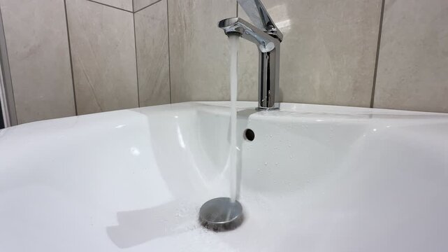 a hand turning on a chrome faucet in a modern white bathroom sink. Clear water runs steadily down the drain against a tiled wall background, depicting daily home hygiene routines.