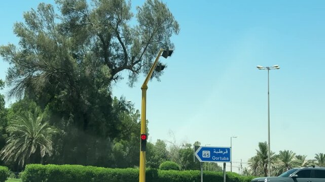 Wide shot of cars waiting at a red traffic light on a sunny day in Kuwait. A blue road sign directs traffic toward Qortuba on Route 45, surrounded by green trees and a clear blue sky.