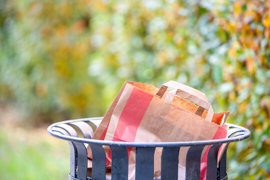 Craft paper bag with handle lying in a metal street trash bin against a blurred green park foliage background, recycling and eco friendly waste disposal concept.