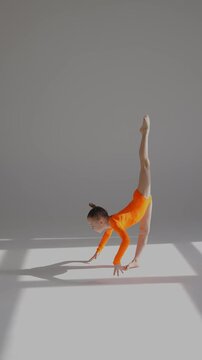 A little Caucasian girl gymnast in an orange leotard demonstrates her skills on a white background. 
