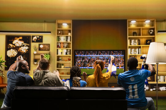 Football supporters sit on sofa to follow live soccer match on large TV. Seen from behind, fans in cozy living room watching soccer broadcast on television screen with enthusiastic body language.