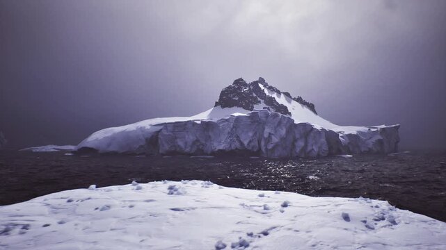 Foreground glacier vista with jagged ice ledge framing turbulent dark sea and distant mountain under heavy clouds, scientific monitoring and coastal survey mood