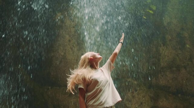 A blonde woman raises her hands to the sky, looking joyfully at a powerful waterfall cascading down in a natural setting. She stands immersed in the water spray.