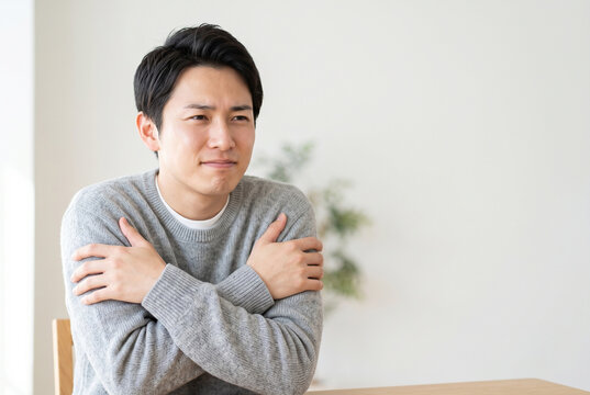 室内で腕を抱え込み寒そうにしている若い男性 Generative AI