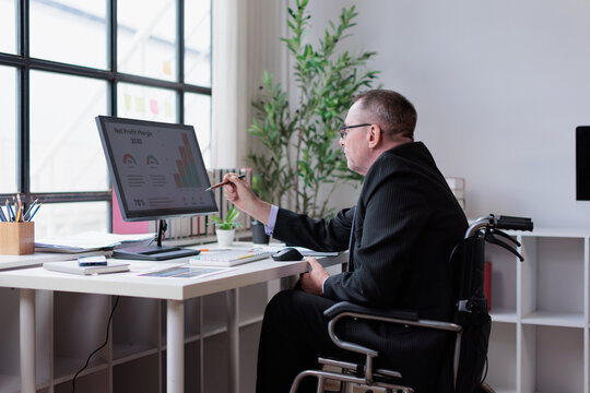 Business professional in wheelchair analyzing financial data on computer monitor, working in modern office, showing diversity and inclusion in workforce