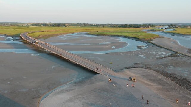 Drone view of the causeway leading to Mont Saint-Michel, revealing the tidal flats, winding channels, and visitors crossing the iconic access bridge in Normandy, France