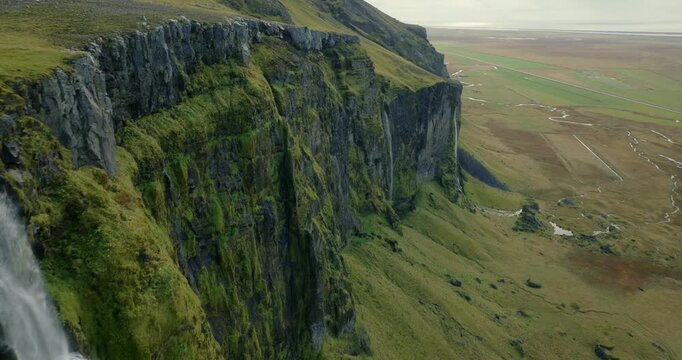 Seljalandsfoss plunging from towering basalt cliffs above the Seljalandsa River in Rangarthing eystra Iceland exposing volcanic escarpments, moss-covered rocks and coastal lowlands, drone forward fly