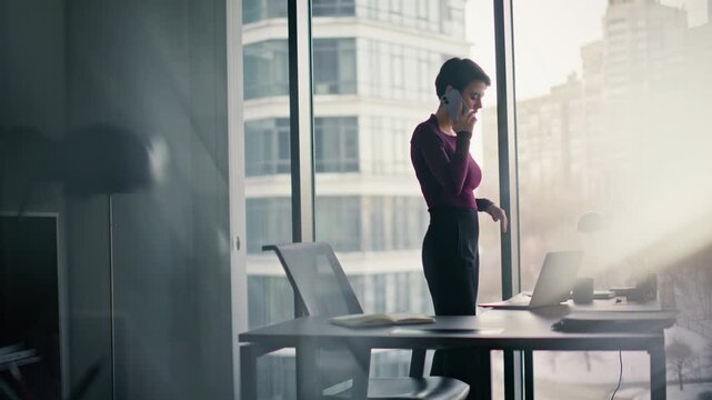 Businesswoman phone call working laptop office. Pensive lady standing window.
