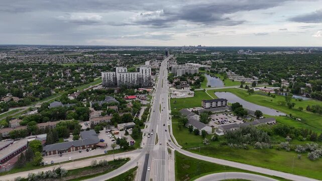 Drone flying high above Pembina Highway in Winnipeg, backing up to reveal the Perimeter Highway interchange near St. Norbert with cars, cloverleaf ramps, suburbs, green grass and partly cloudy summer