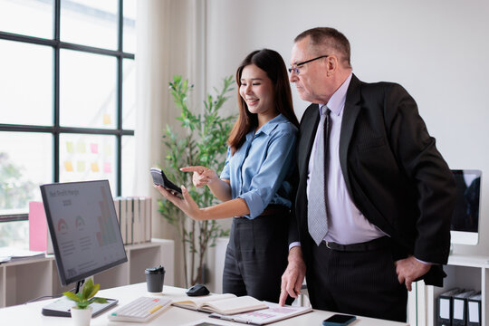 Business manager mentoring young businesswoman showing financial data on smartphone, discussing results during teamwork in modern office, analyzing company profits and strategies