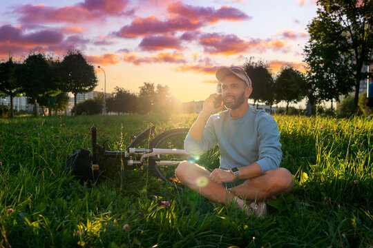 Smiling guy relaxes on the green meadow in a park at sunrise while having a pleasant phone conversation next to his parked mountain bike outdoors