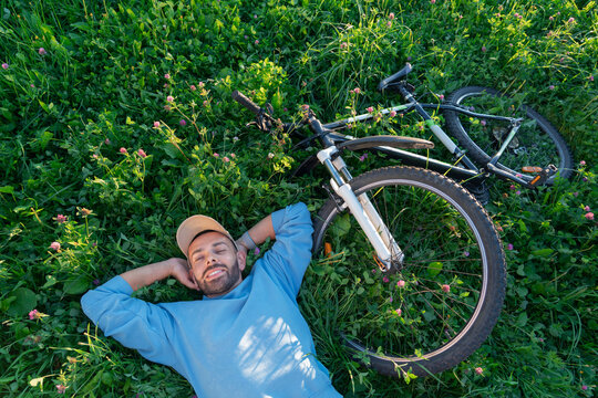 Smiling guy in a blue sweater relaxing on the meadow near his bike during a sunny summer morning to enjoy peace and rest in a beautiful city park