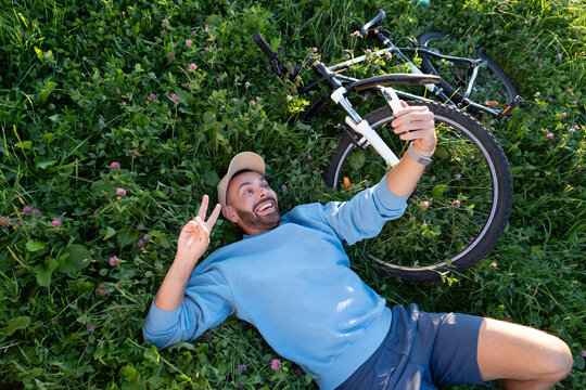 Cheerful man relaxes on the grass in a city park during a summer morning, smiling and gesturing for a selfie with his phone next to a parked bicycle