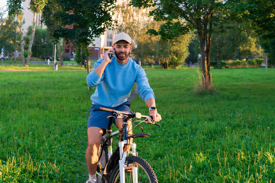 Happy man wearing a baseball cap enjoys a summer morning ride on his bicycle in a city park while talking on his phone and smiling at the camera lens