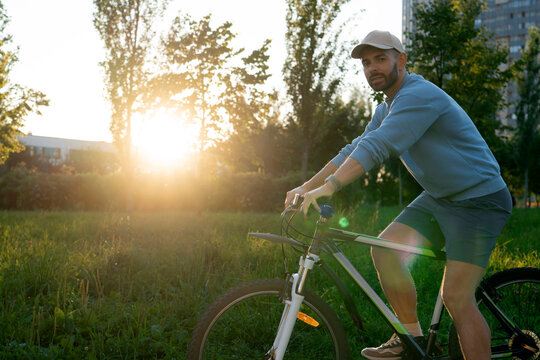 Young bearded man wearing a cap rides his bicycle across a sunlit grassy meadow in the park on a bright summer morning while looking toward the camera