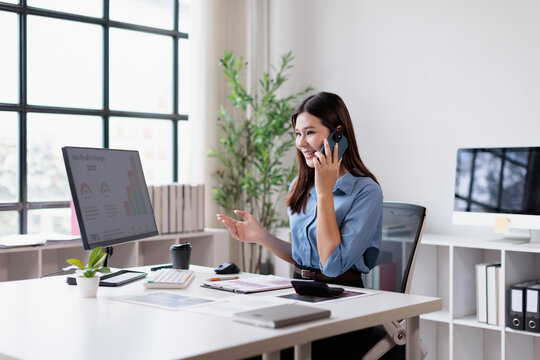 Young asian businesswoman actively communicating on her mobile phone. Managing business tasks. Analyzing financial reports on a computer