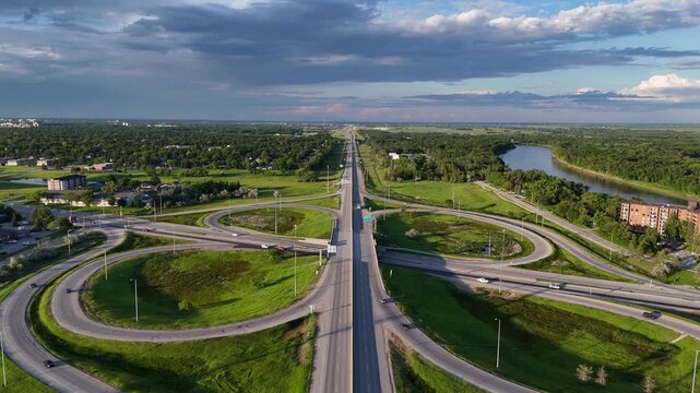 Drone aerial of the Perimeter Highway cloverleaf interchange in St. Norbert, Winnipeg, Manitoba, Canada. Camera slowly backs up east along highway showing traffic, Red River, grass, buildings.
