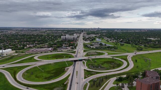 Drone flying forward above Pembina Highway in Winnipeg, revealing the Perimeter Highway interchange near St. Norbert with cars, cloverleaf ramps, suburbs, green grass and partly cloudy summer skies.