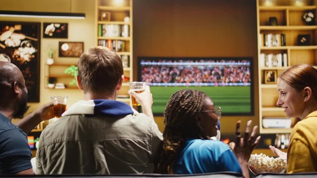 Soccer supporters eat popcorn on the sofa during a home match watch party while football fans enjoy snacks and react to the live game on TV.