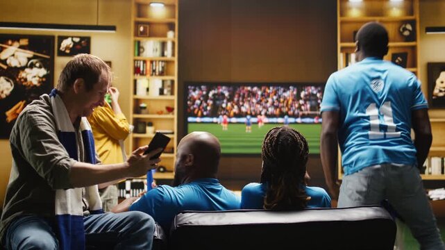A delighted soccer fan sees a positive match result on a smartphone screen, showing a friend good news and sharing excitement at home.