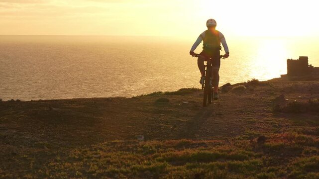 Mountain biker silhouette riding a bicycle on a cliffside trail over the ocean. Golden hour sunlight creating a stunning, warm light