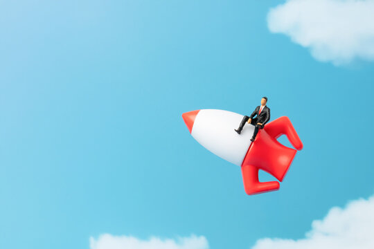 Businessman figurine sitting on a rocket ship flying upwards through a blue sky with clouds. Startup launch, career growth, acceleration, ambition concept.