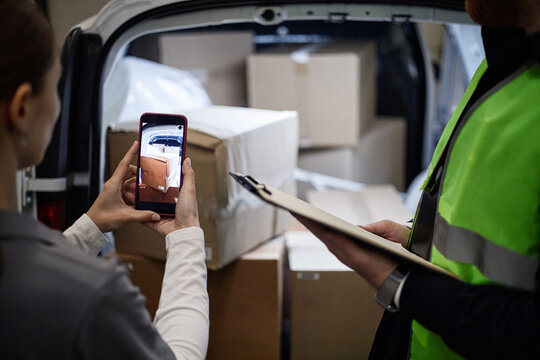 Young adult woman photographing packed boxes in vehicle while delivery worker checking clipboard, documenting individual storage inventory. Useful for logistics, moving, self storage marketing
