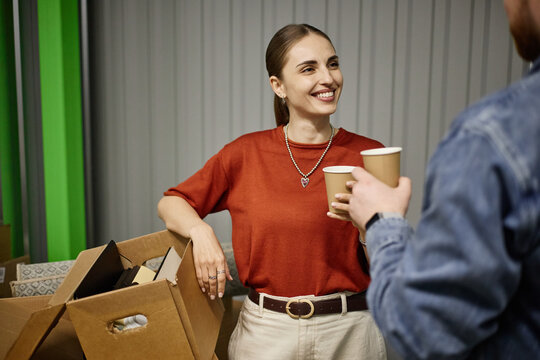 Young adult Caucasian woman discussing individual storage rental with man while holding coffee near packed boxes, suggesting moving or decluttering service. Medium shot for self storage marketing