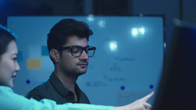 Young man with eyeglasses looking at monitor during discussion with colleague near whiteboard, software team meeting in dark office.