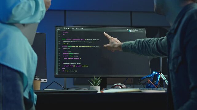 Man pointing at screen with colorful source code while colleague with laptop watches, pair programming and debugging in dark blue office.