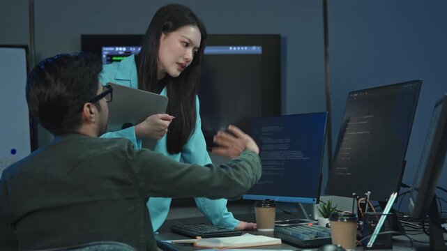 Male Programmer pointing at monitor with program code as Asian woman holding tablet looks on, problem solving teamwork in office.