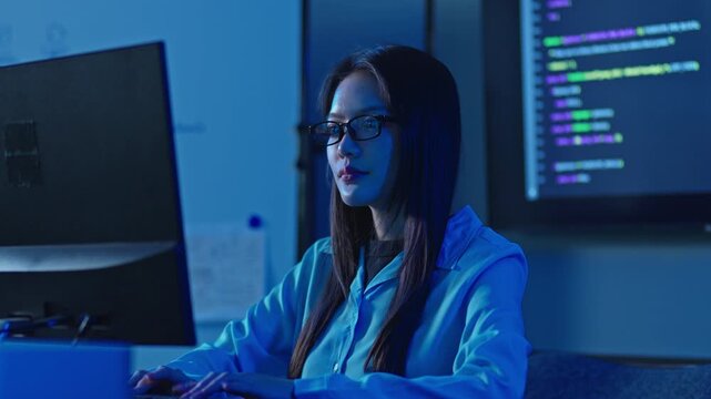 Asian woman with eyeglasses working at computer with program code on screen beside her, focused software engineer typing late at night.