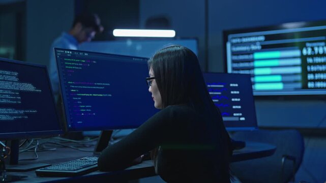 Asian woman with glasses sitting at multiple monitors with program code and statistics, male colleague behind, dark monitoring room.