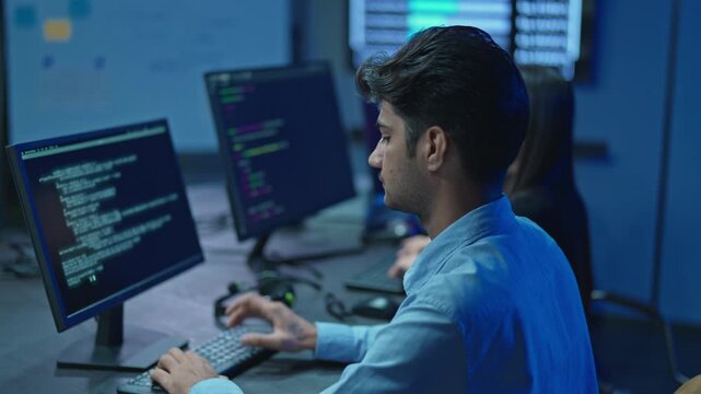 Male Programmer at desk with program code on screens, female colleague in background, software engineering team working late in blue light.