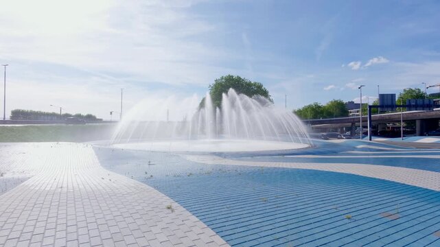 Large modern fountain spraying water upwards and outwards in a public square with unique blue and white paving patterns. Busy highway traffic visible in the background under a clear summer sky.