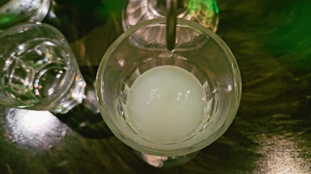 Detailed overhead shot of absinthe louche in a glass, showing pale cloudy drink, dripping water spout, dark bar table reflections, and moody nightlife cocktail lounge setting.